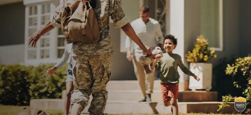 A soldier in uniform returns home, reaching out with a teddy bear. A smiling boy runs towards him, with another adult in the background
