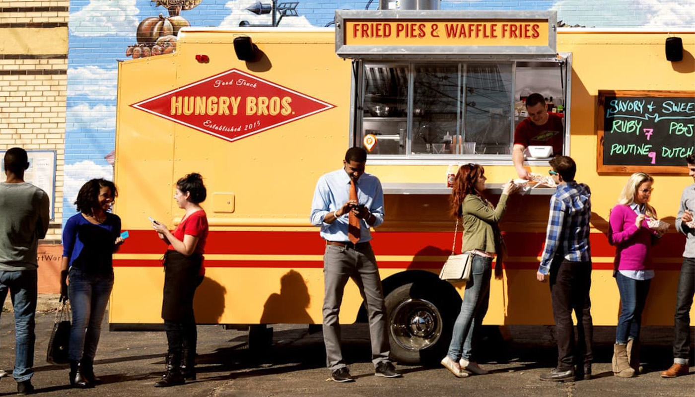 Yellow and red “Hungry Bros.” truck by brick building; truck says “Fried Pies & Waffle Fries”; customers gather and order.