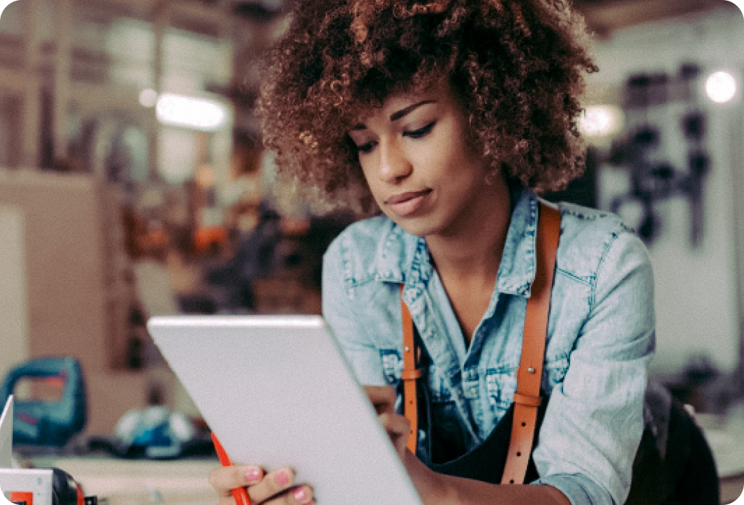 Woman in a woodworking shop wearing an apron on, leaning on a wood table, while working on a tablet