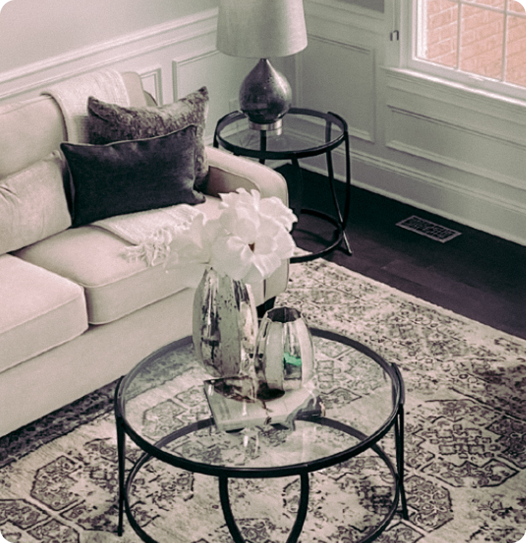 Staged living room with a couch that has blankets and pillows, a glass coffee table sitting on top of a patterned rug, and a glass side table with a lamp on it