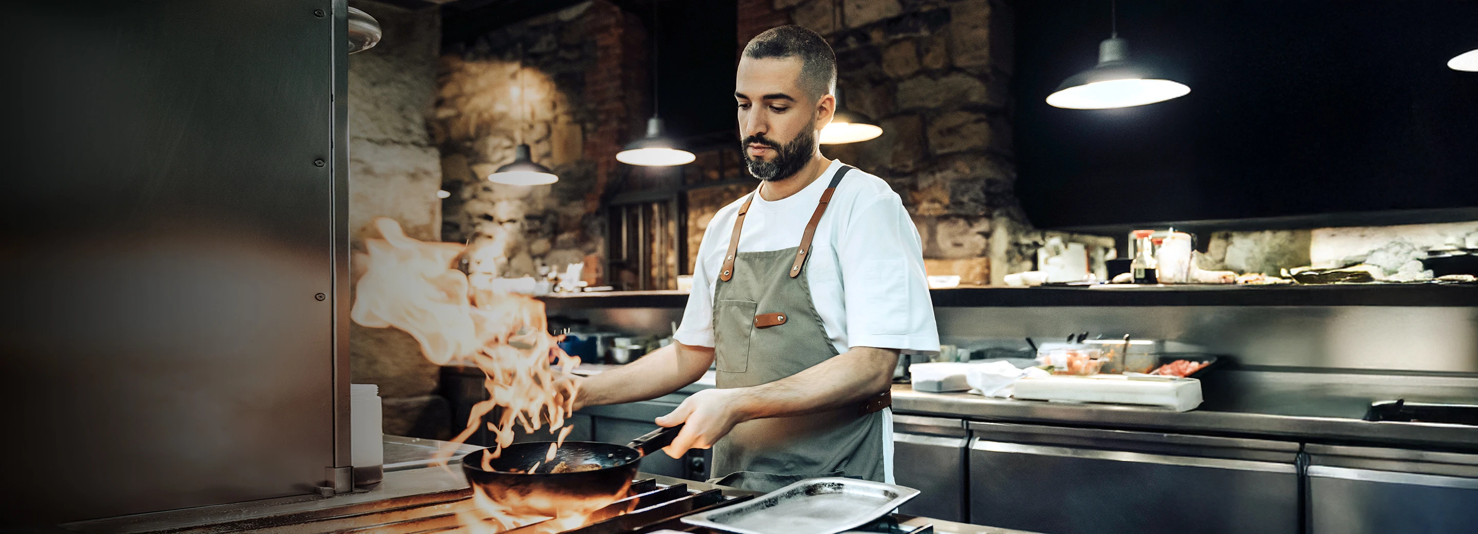 Chef in a kitchen wearing a dark green apron while cooking food on a high flame. A large flame peeks out from under the pan.