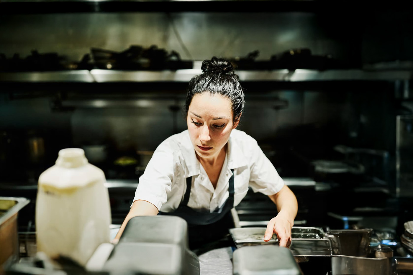 Chef in white shirt and apron works in commercial kitchen; food prep containers and stove visible in clean, focused setting.