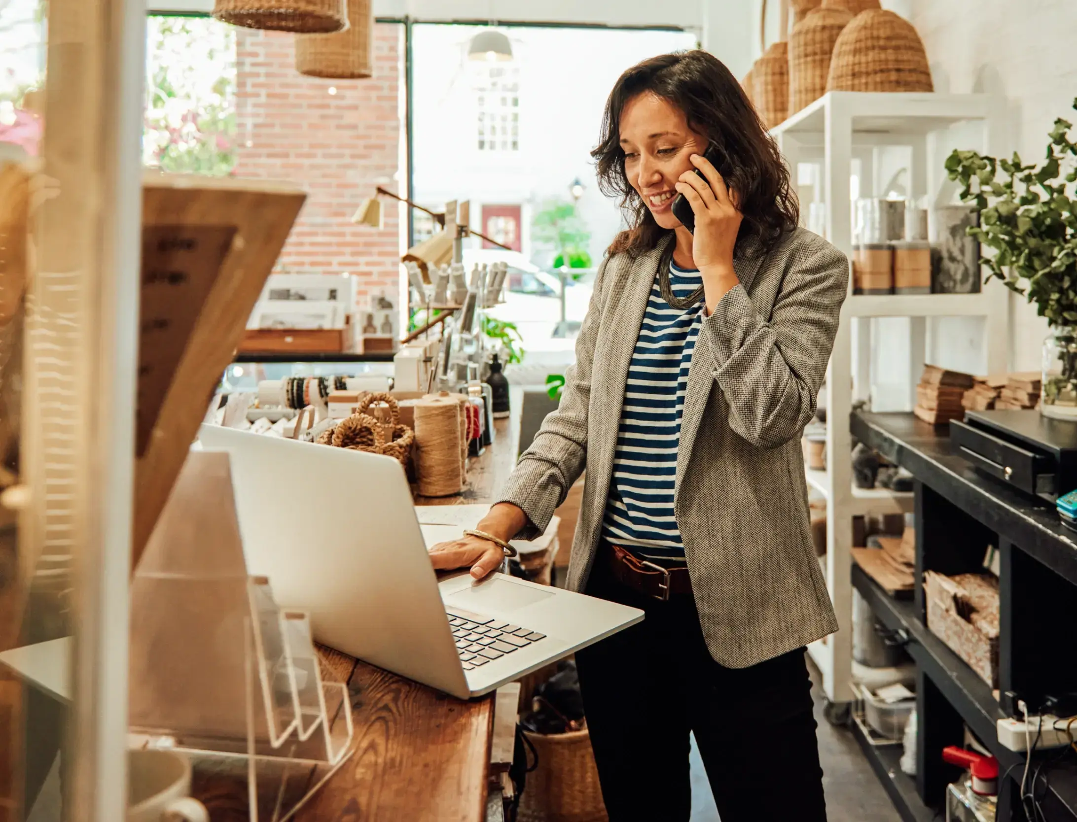 Sales woman dressed in business attire standing behind a counter talking on phone while using a laptop.