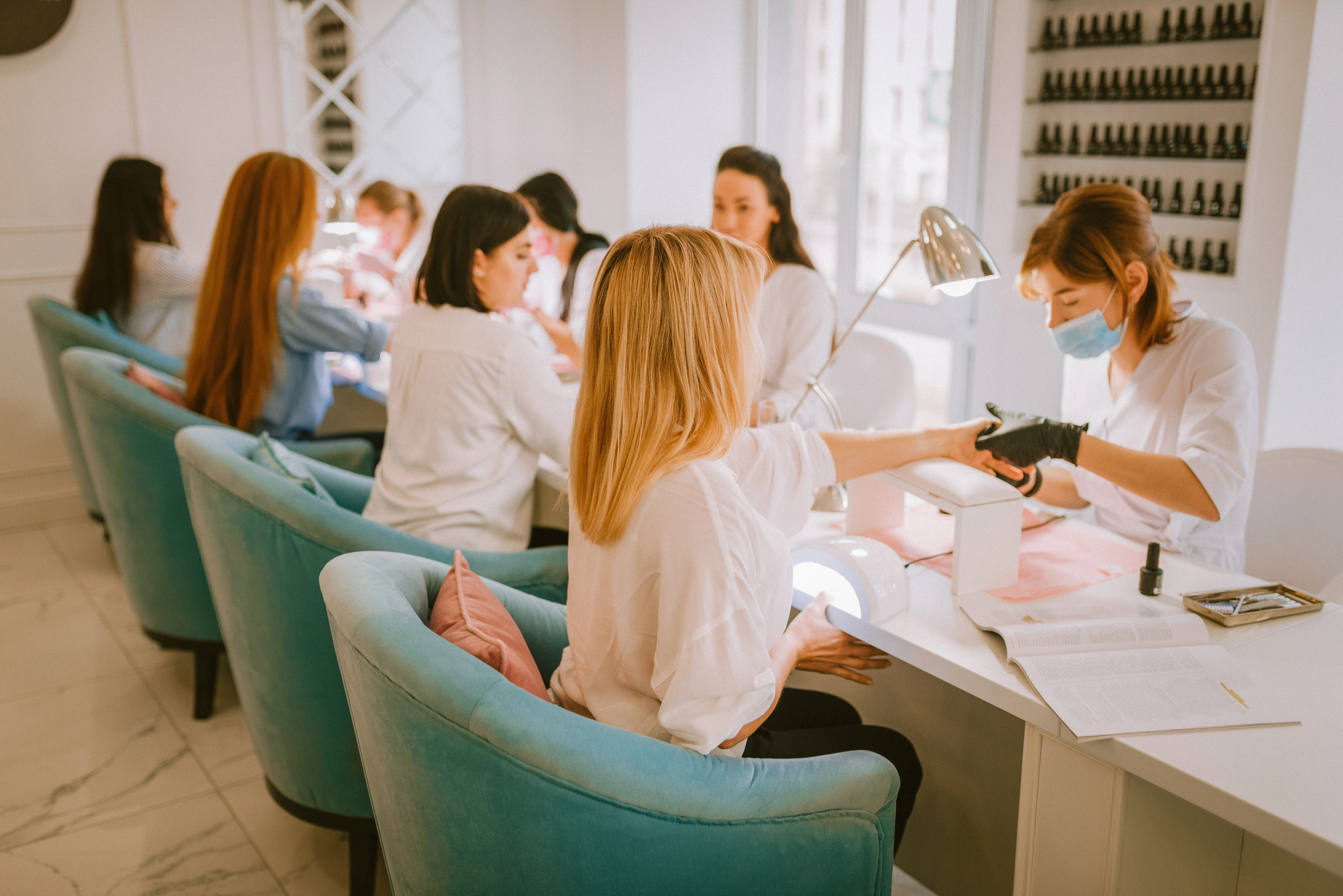 Four women sitting in a row in turquoise chairs at a nail salon getting their nails done. Nail techs sit across from them.