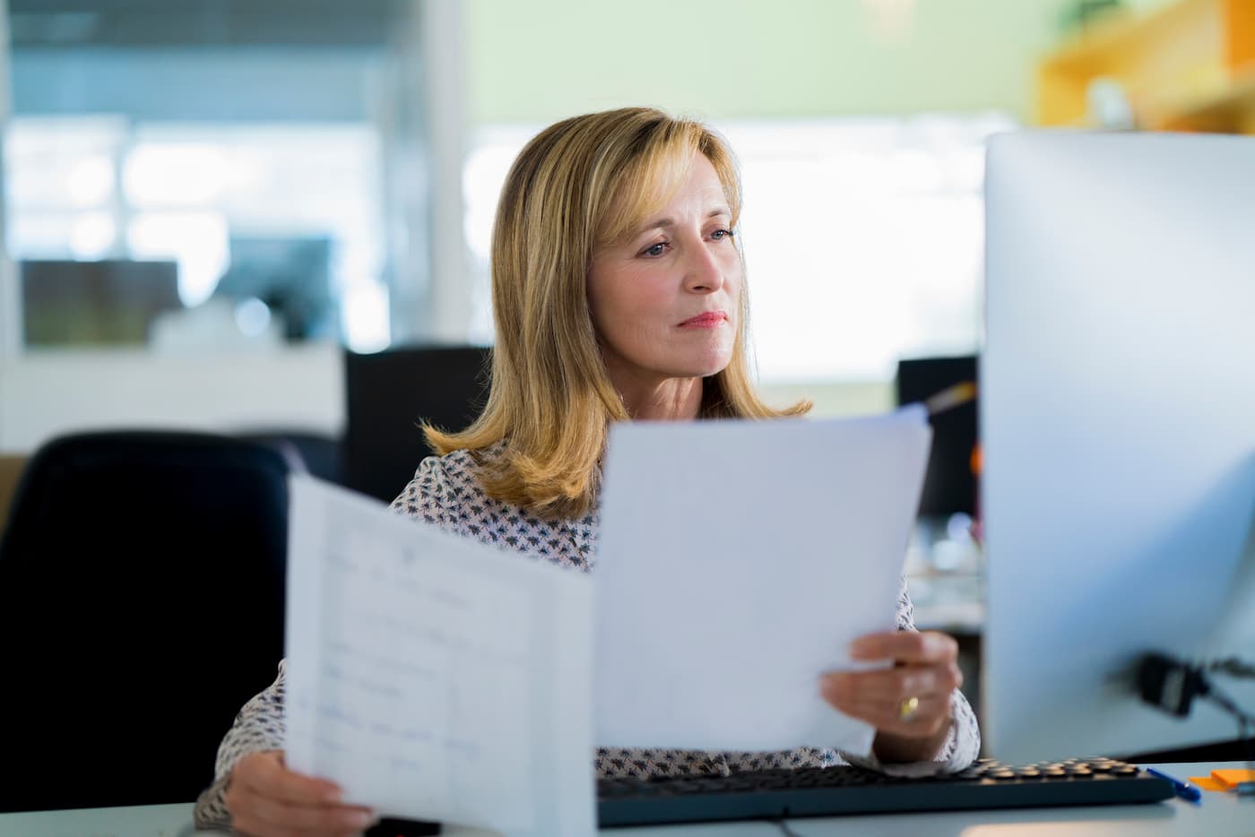 A professional woman holding papers in both her hands and looking at monitor.