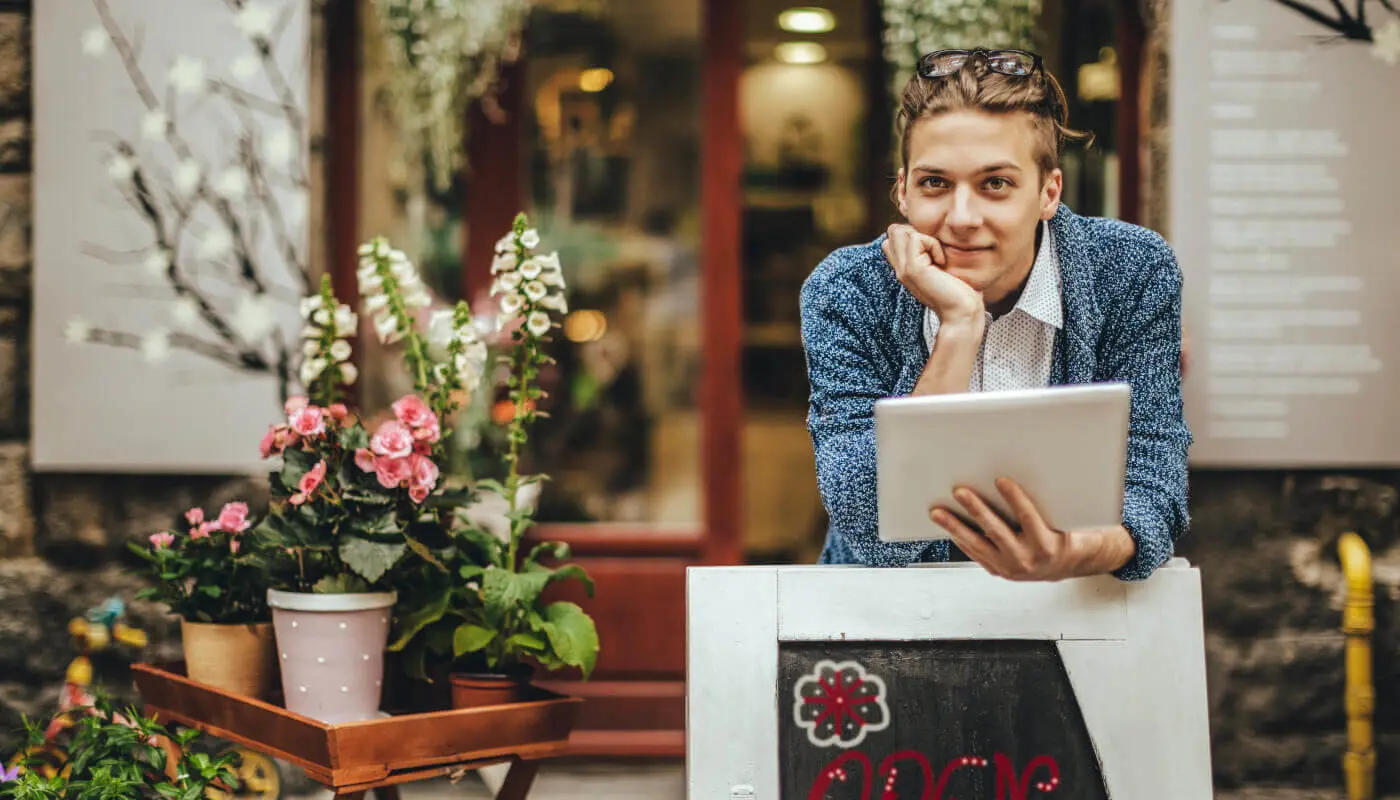 Person standing outside a flower shop leaning over a wood sidewalk sign while holding a tablet and gazing into the distance.