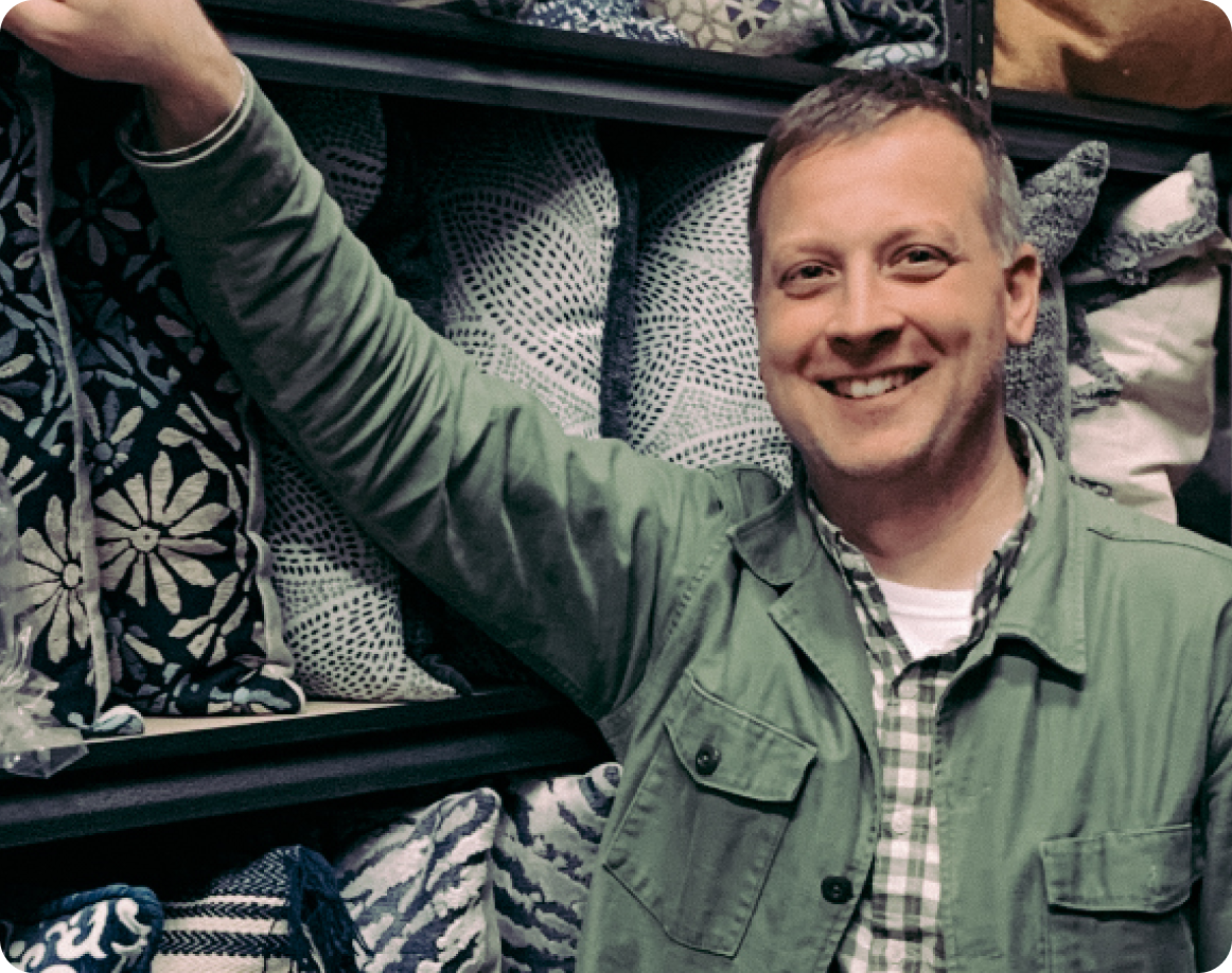 Owner of Fox Design and Staging company smiling, standing in front of a rack of pillows with his arm resting against the rack