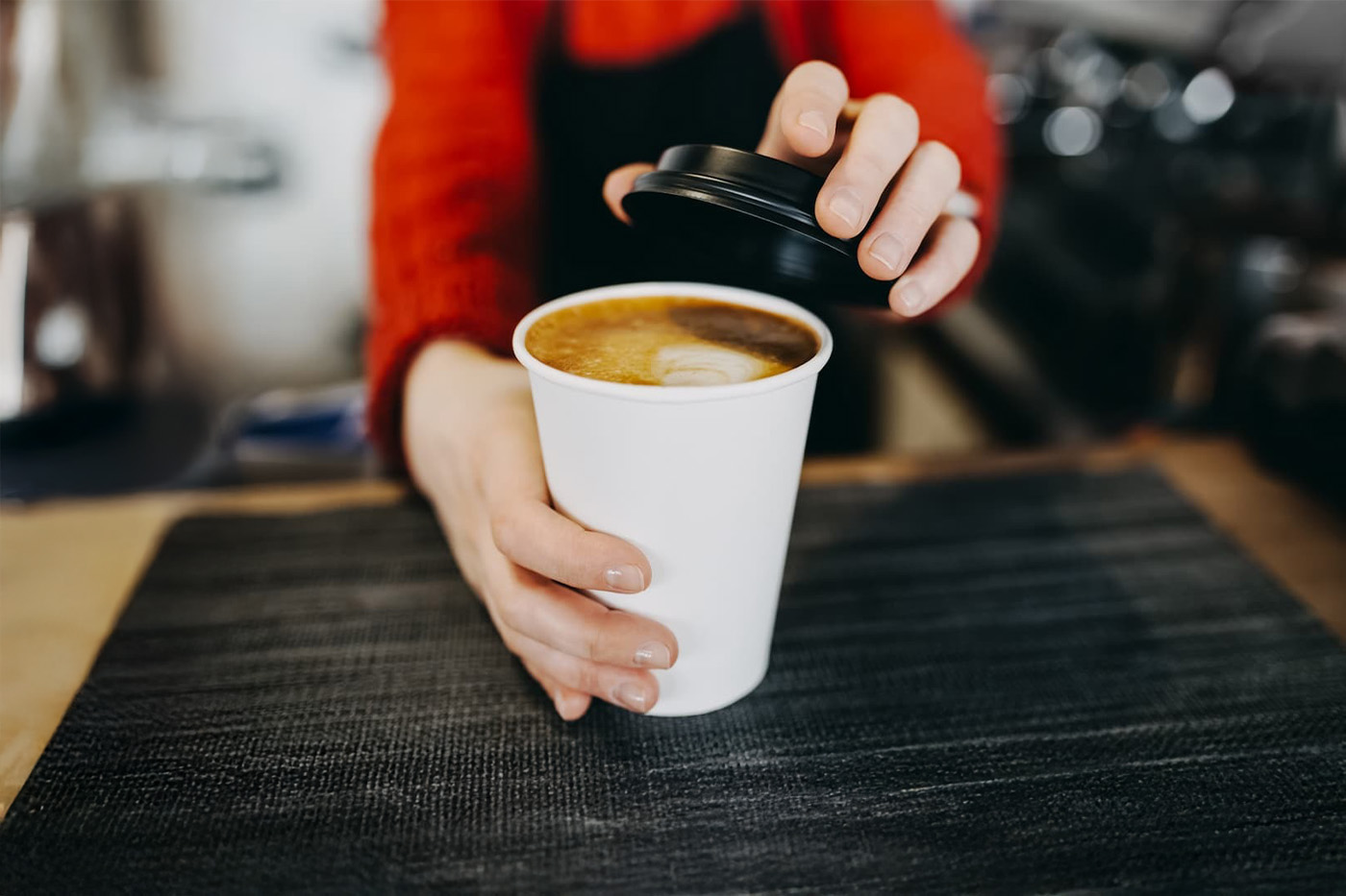 Barista wearing red sweater and apron places lid on latte with foam art; close-up of hands, blurred café background.