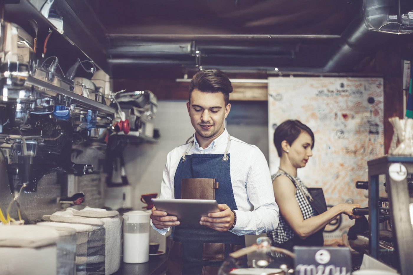 Male barista in white shirt and blue apron uses tablet; female barista makes espresso in back. Barista area in view.