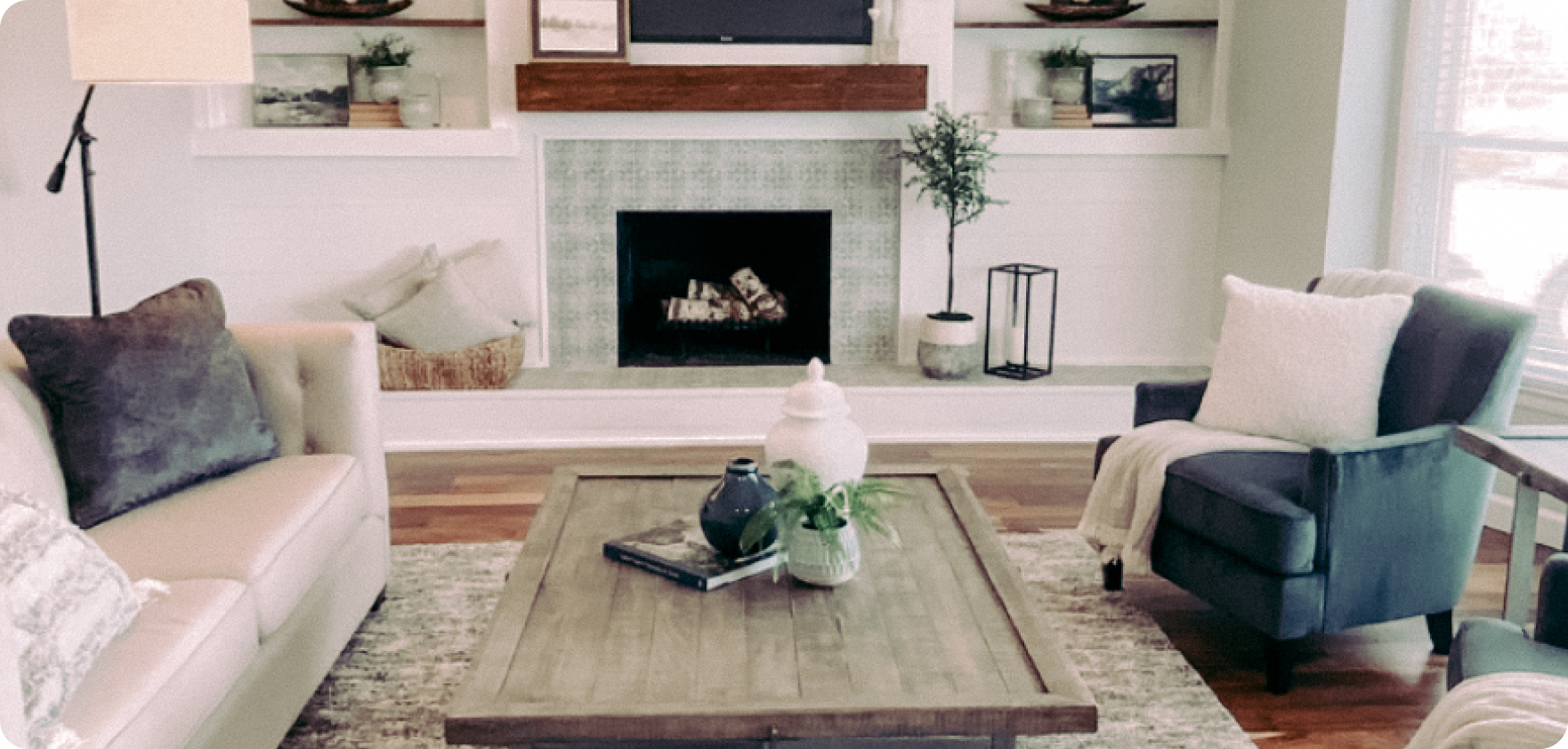 Staged living room that has a fireplace, bookshelves, a beige couch with pillows, a wood table with decorations, and two armchairs with pillows and blankets