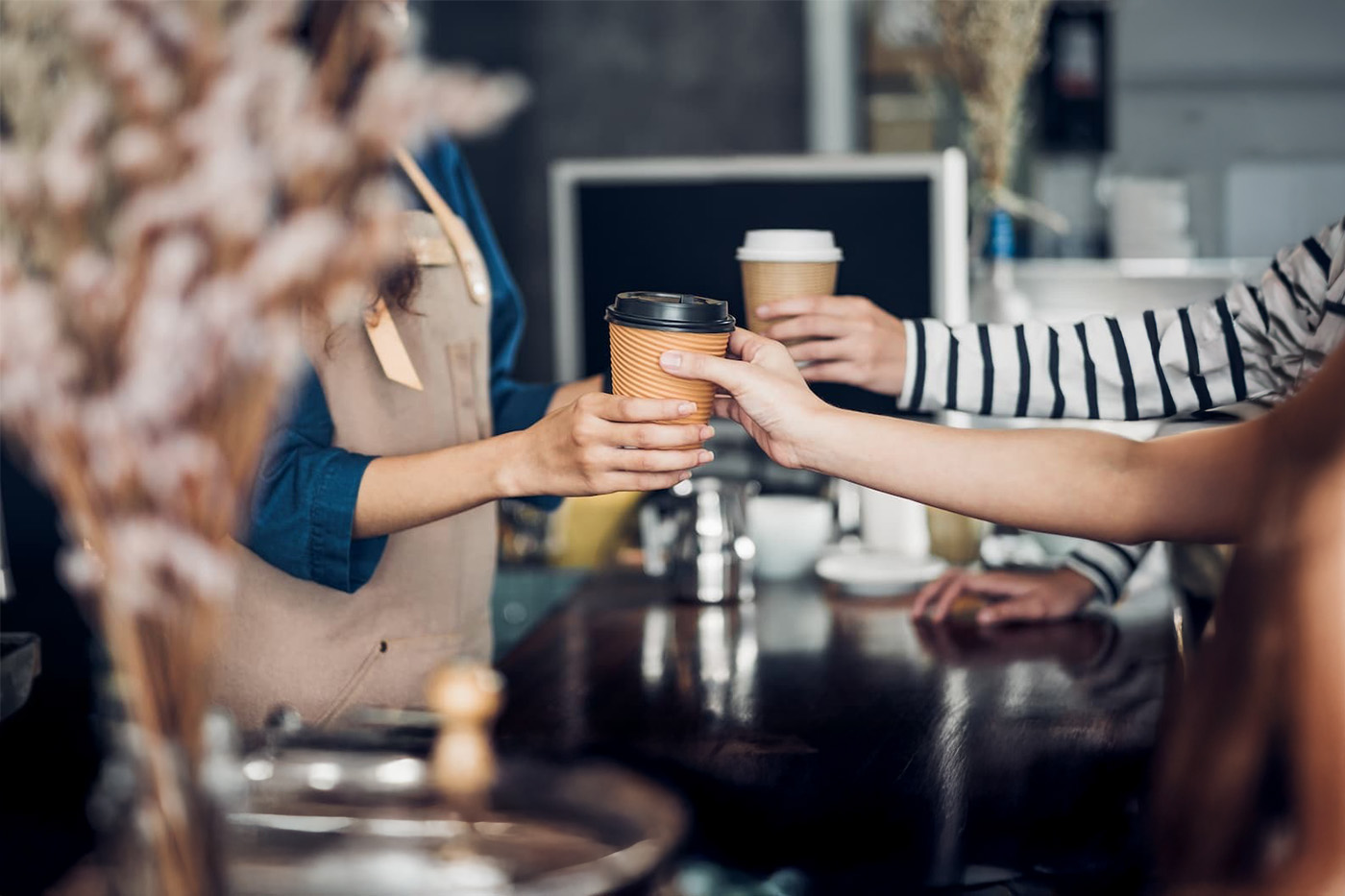 Barista in beige apron hands coffee to customer at café counter; another customer in striped shirt reaches holding coffee.