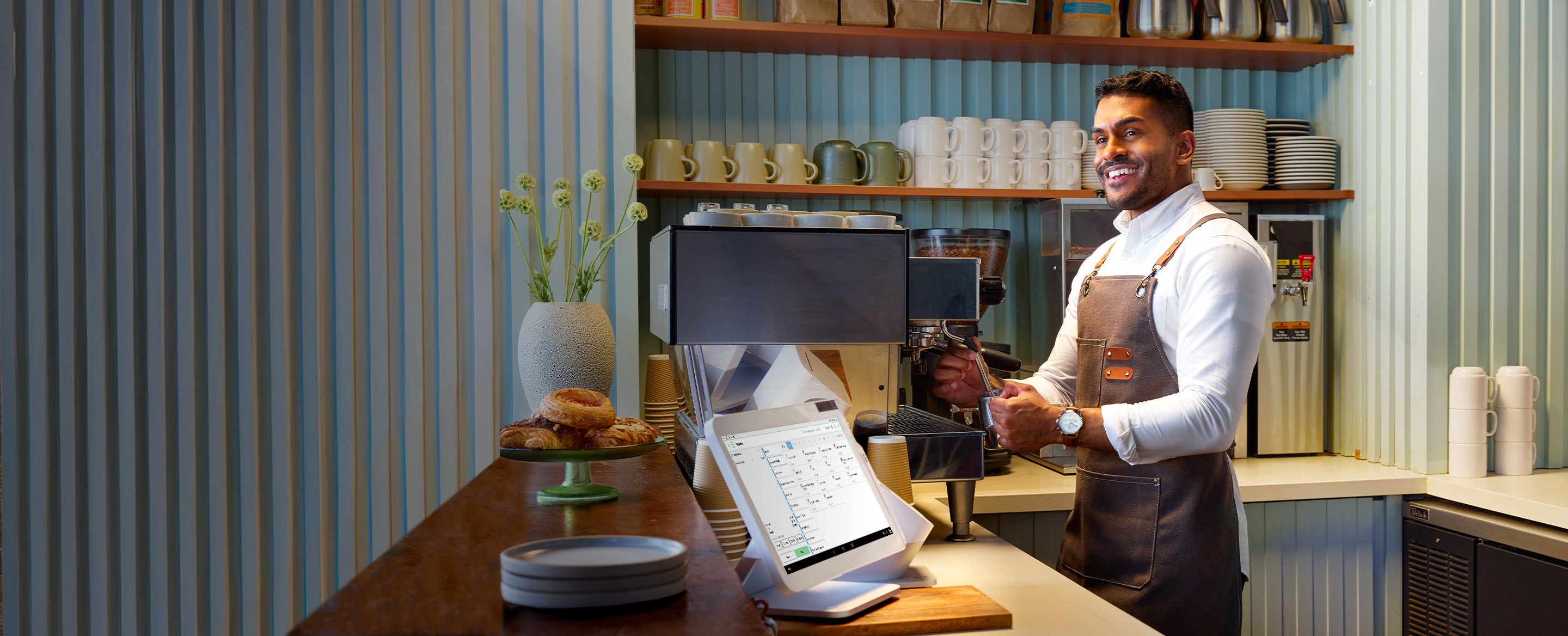 Barista in white shirt and brown apron smiles while using espresso machine; doughnuts, clover station solo, and mugs visible.