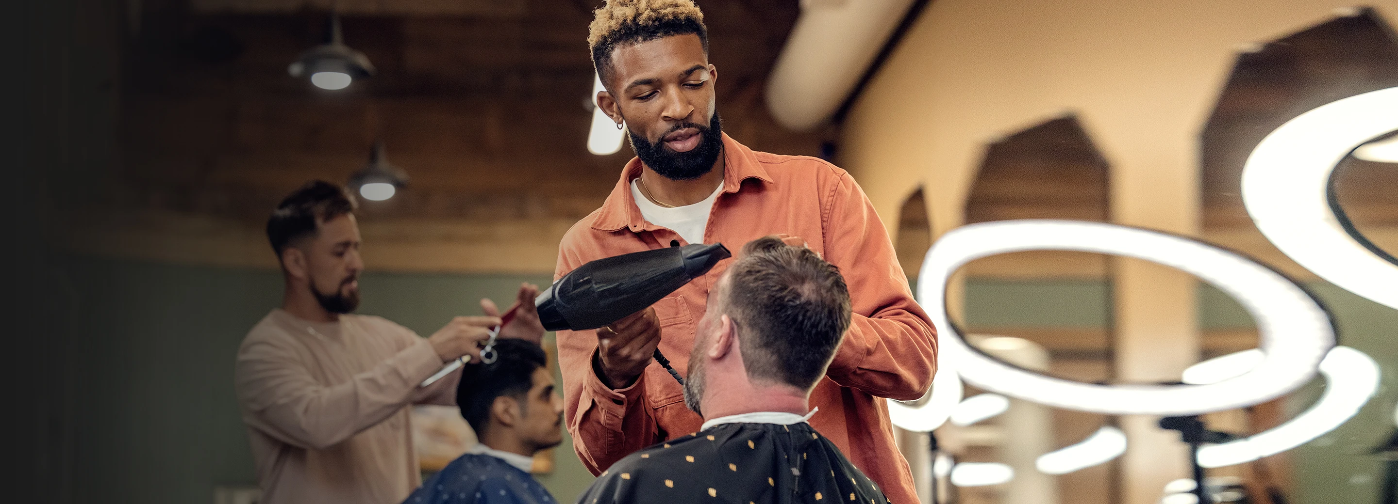 Male barber styling a male customer's hair with a blow dryer while he sits in a chair with an apron over his body.