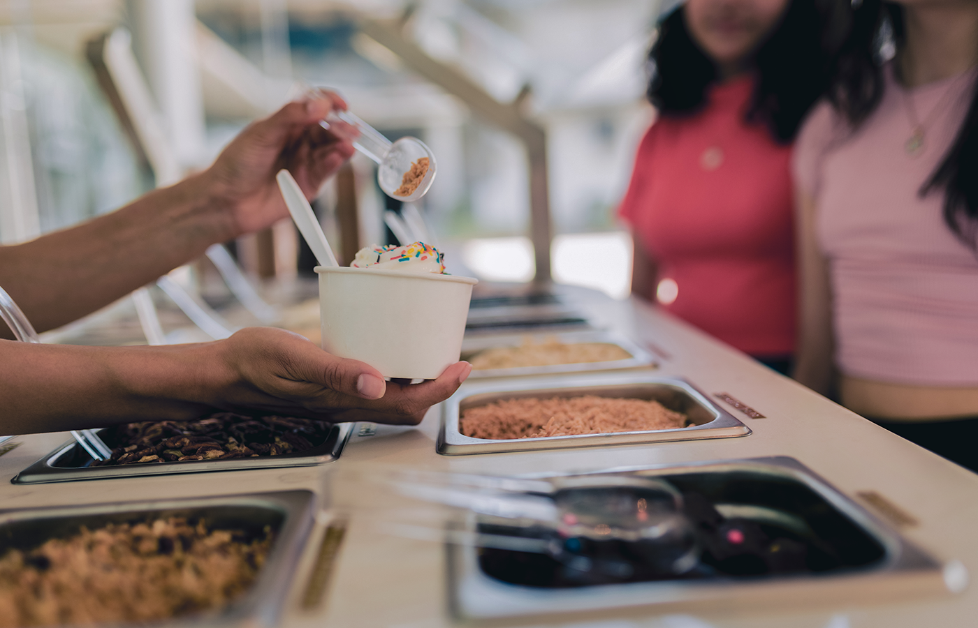 Store attendant pours toppings on a cup of frozen yogurt while customers wait for order