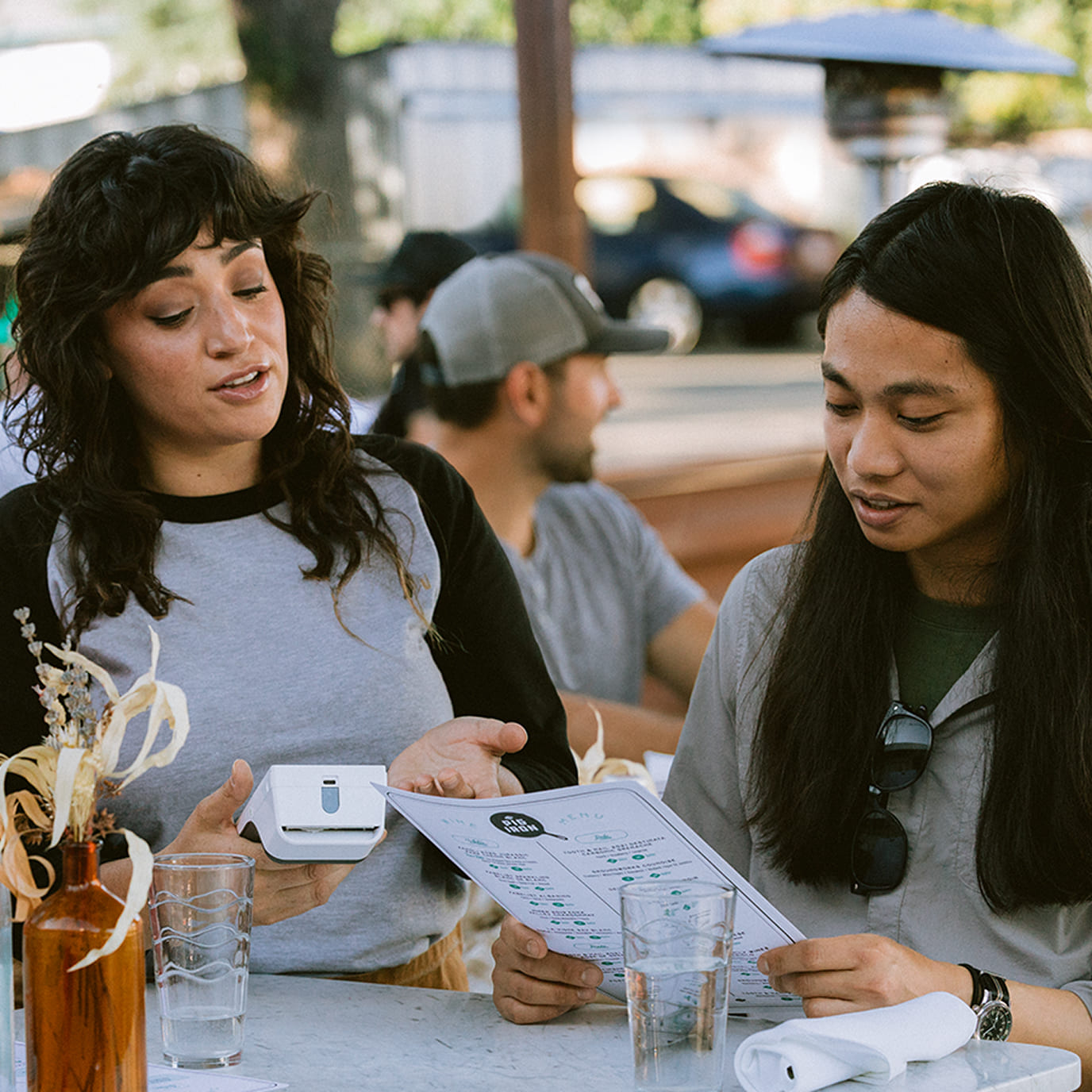 A guest is placing an order and a waitress is holding a device to get the order. A man is sitting wearing a cap at the background.