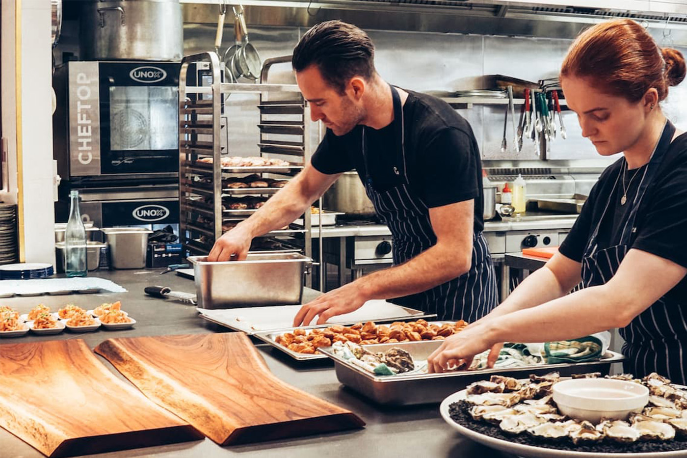 Two chefs in striped aprons prep food at stainless counter; oysters and sauce on serving boards; busy kitchen in background.