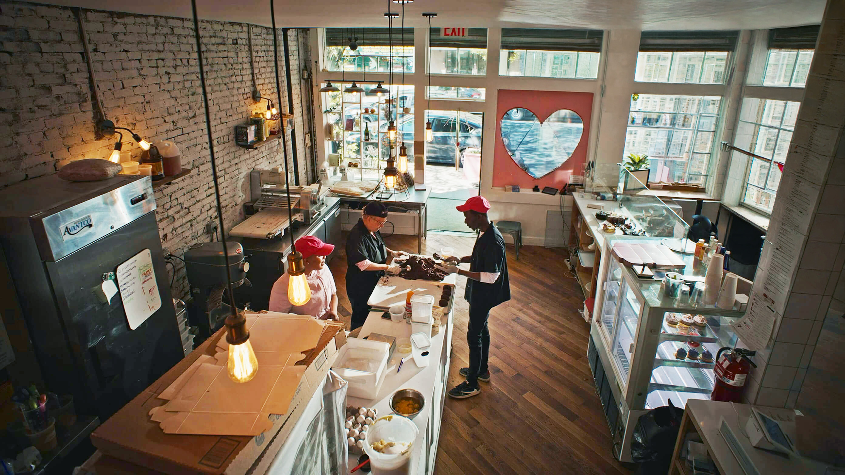 Workers are preparing pastries in a warm, rustic bakery setting; brick wall, heart window, baking equipment visible.