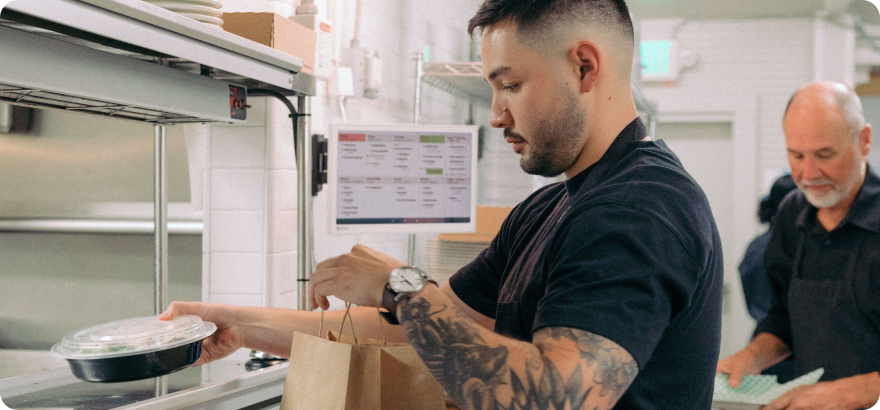 Server putting a take-out container into a paper bag with a Clover Kitchen Display System in the background.