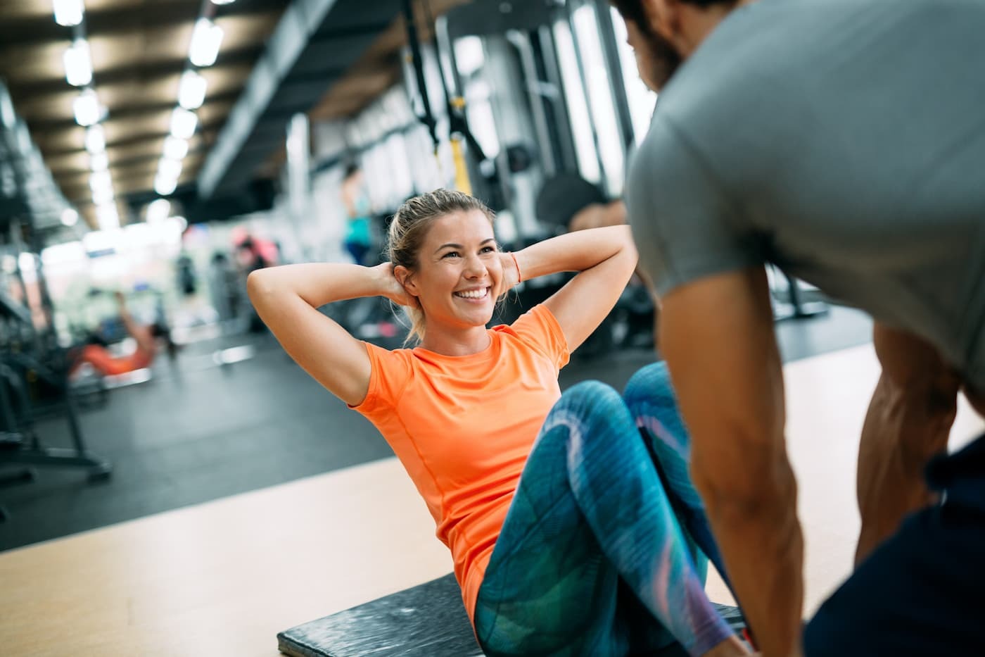 Woman in a gym on a yoga mat in a seated position with her knees bent doing crunches while a male trainer holds her feet.