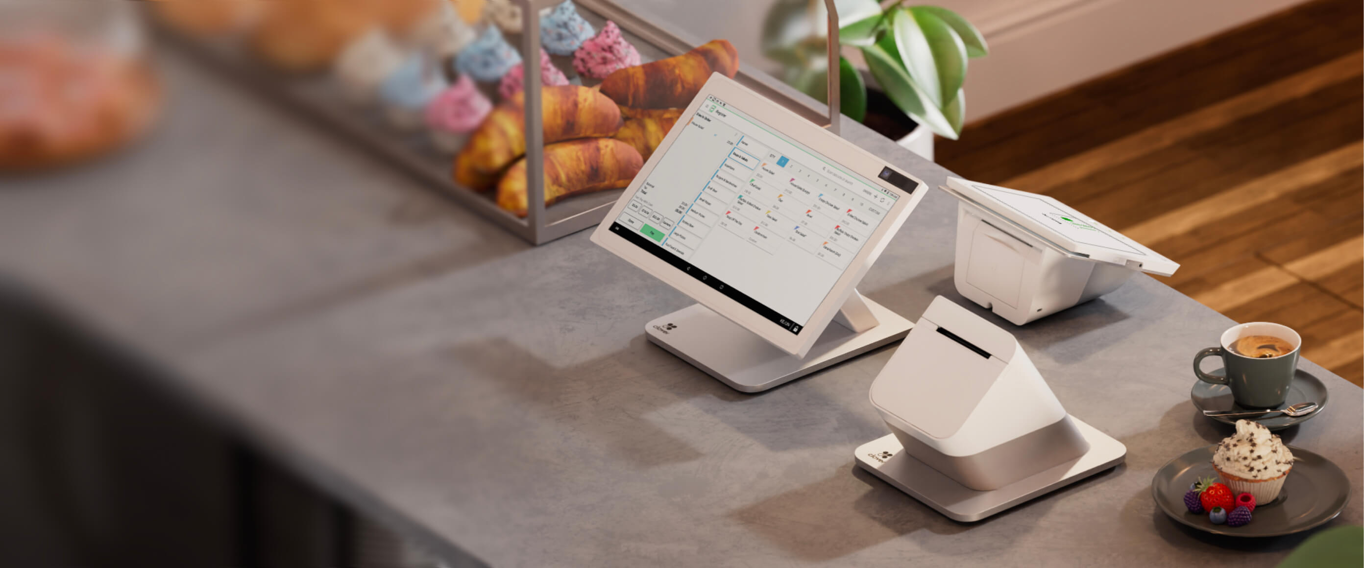 Station Duo on café counter with shop items displayed on screen; blurred pastries, coffee, and dessert in foreground.