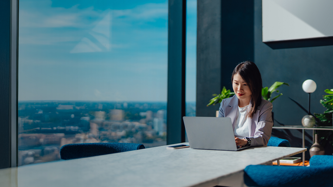 Woman sitting in a modern conference room at a long table working on a laptop. View of a city can be seen out the window.