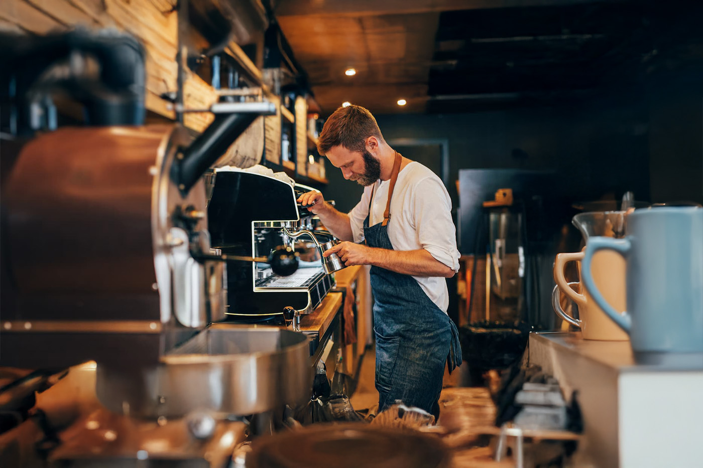 Barista in white shirt and blue apron steams milk at espresso machine; coffee roaster and tools visible in warm café setting.