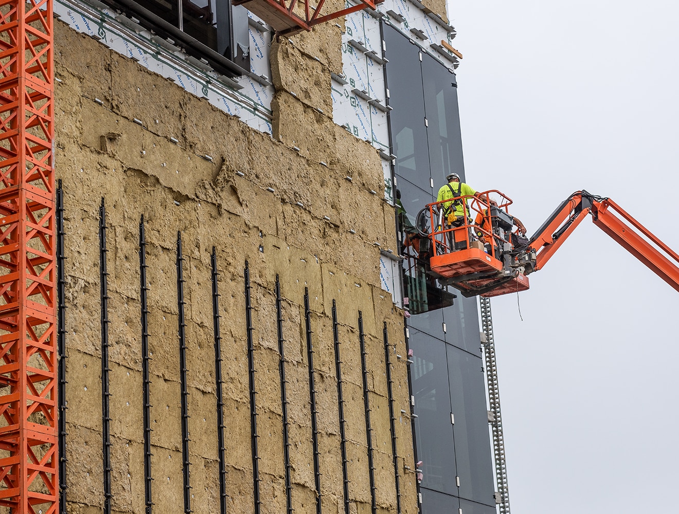 Workers in crane basket inspecting side paneling of building next to exposed insulation material