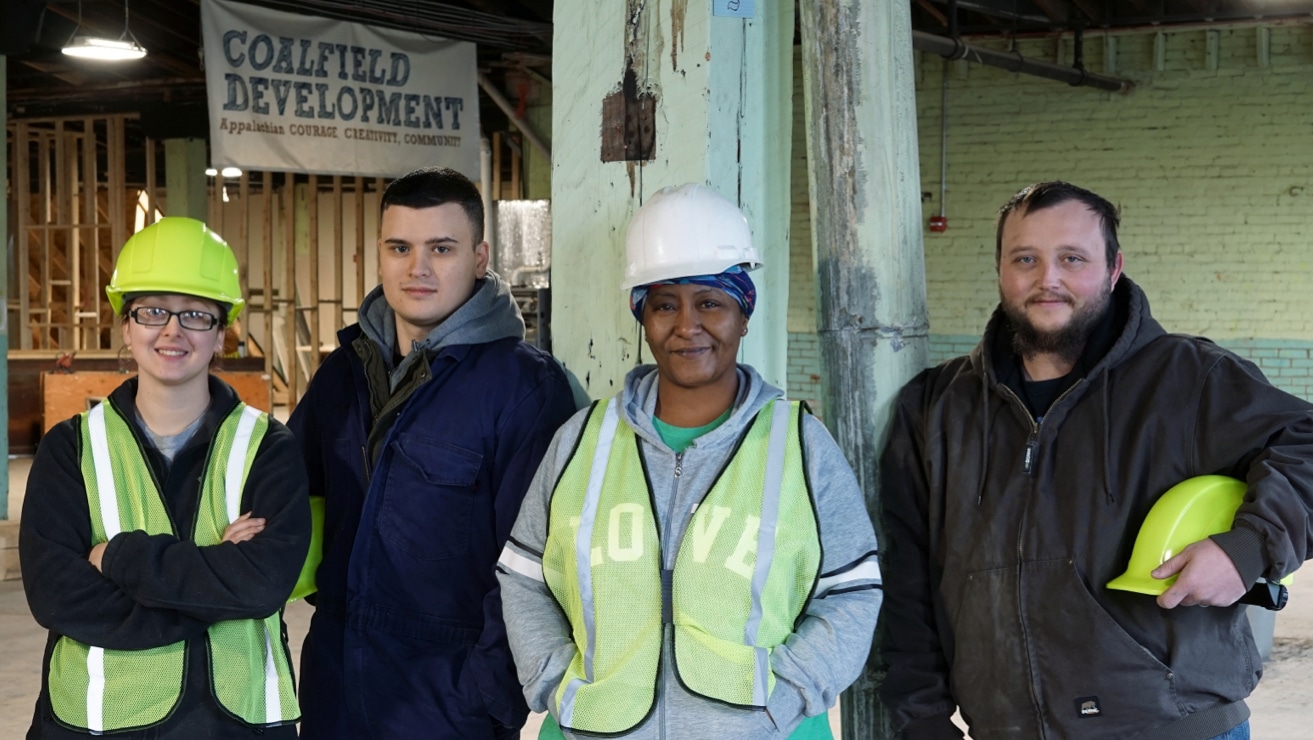 Four Coalfield Development trainees smiling and posing for a group photo — two women wearing hard hats and hi-viz vests, and two men wearing hoodies.