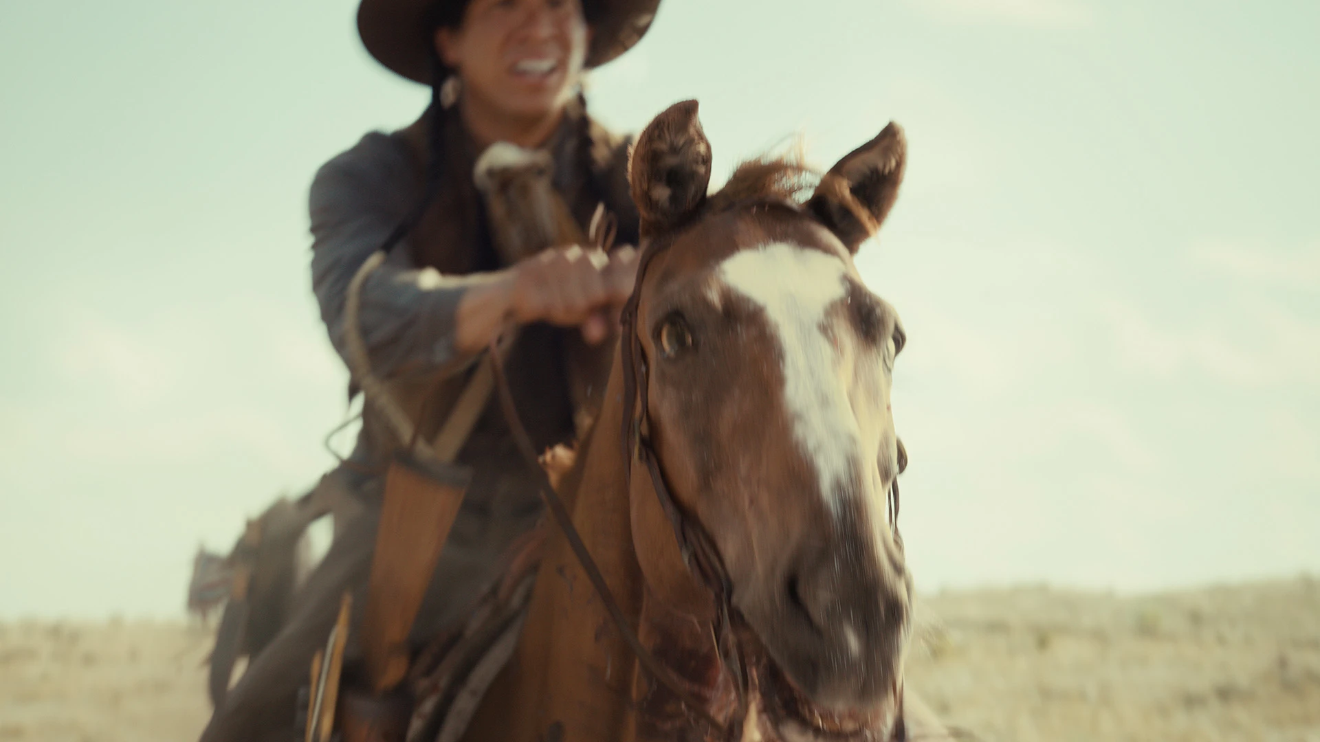 A person wearing a hat rides a brown horse across a sunlit, grassy plain.
