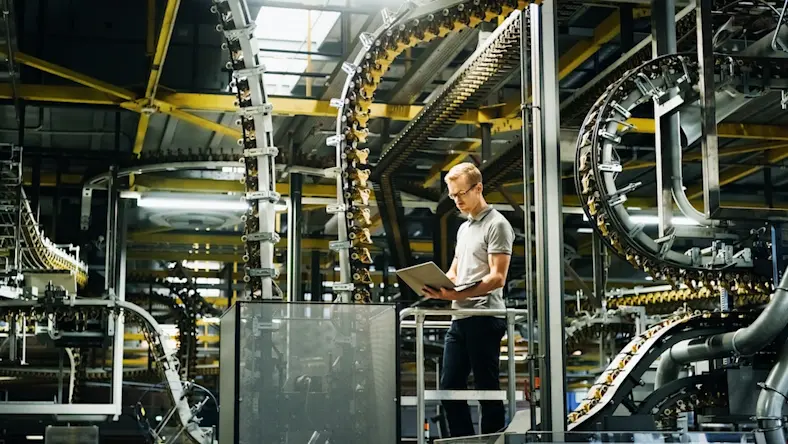 A man in glasses stands looking at laptop in a factory.