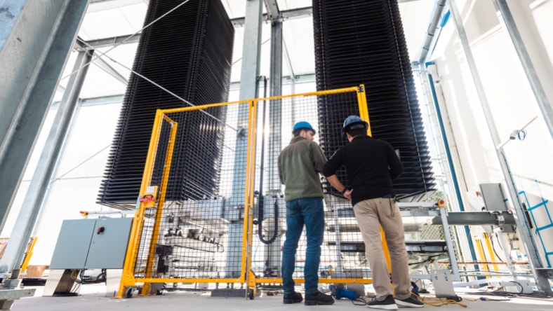 Two Heirloom engineers wearing hardhats, facing away from the camera, standing in front of Heirloom's direct air capture unit — two tall black square columns surrounded by metal scaffolding and caging around the base of the technology. Bright, sunny day.