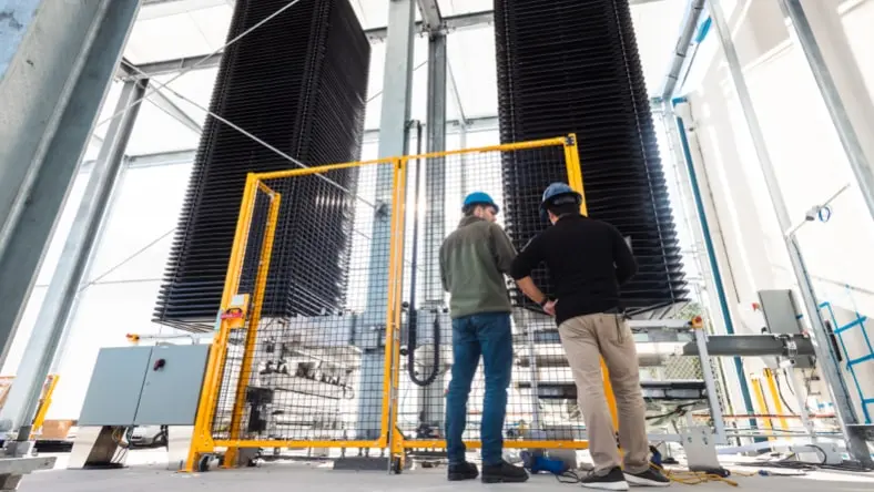 Two Heirloom engineers wearing hardhats, facing away from the camera, standing in front of Heirloom's direct air capture unit — two tall black square columns surrounded by metal scaffolding and caging around the base of the technology. Bright, sunny day.