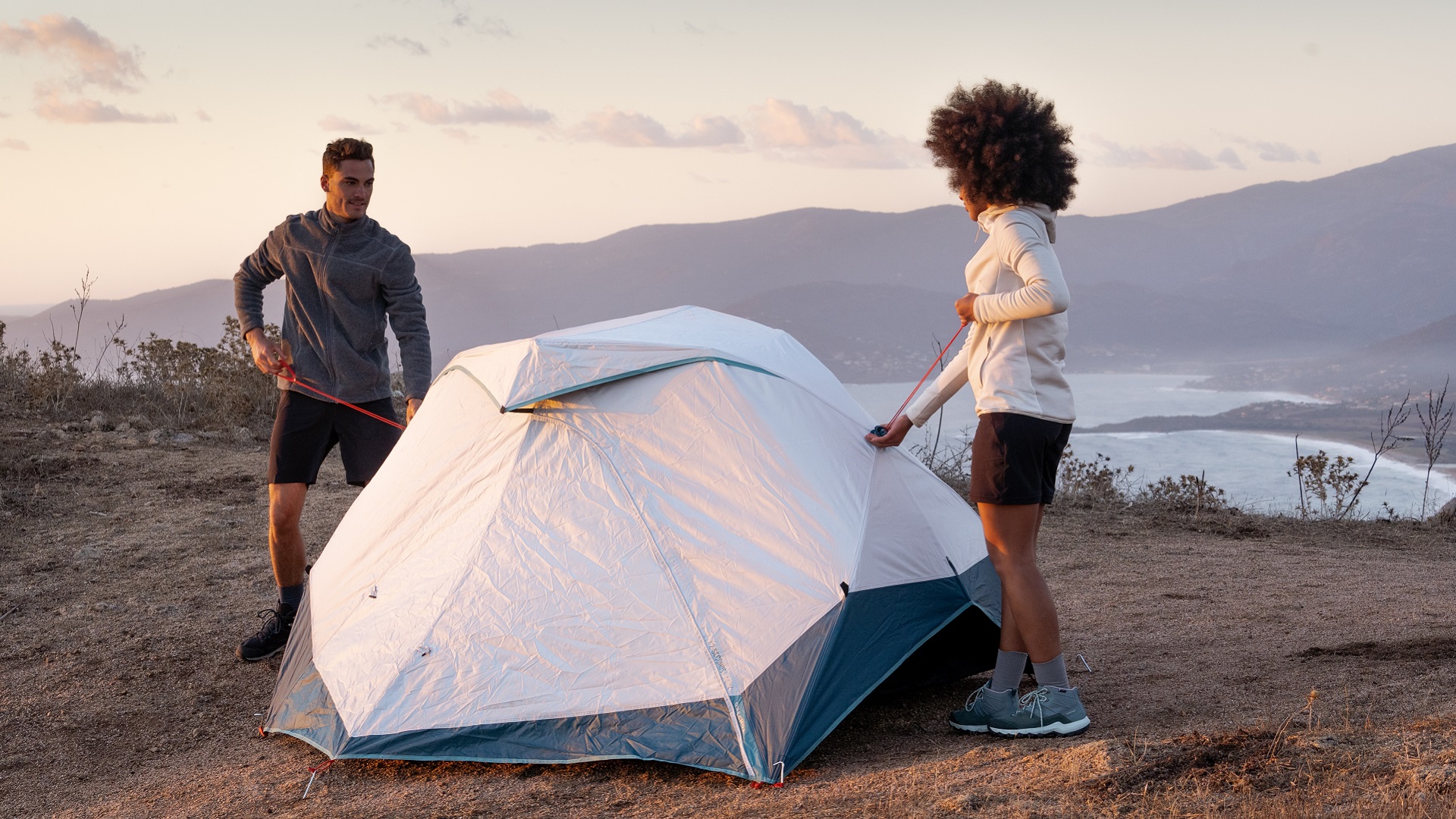 Two people set up a camping tent on a bluff overlooking a lake.