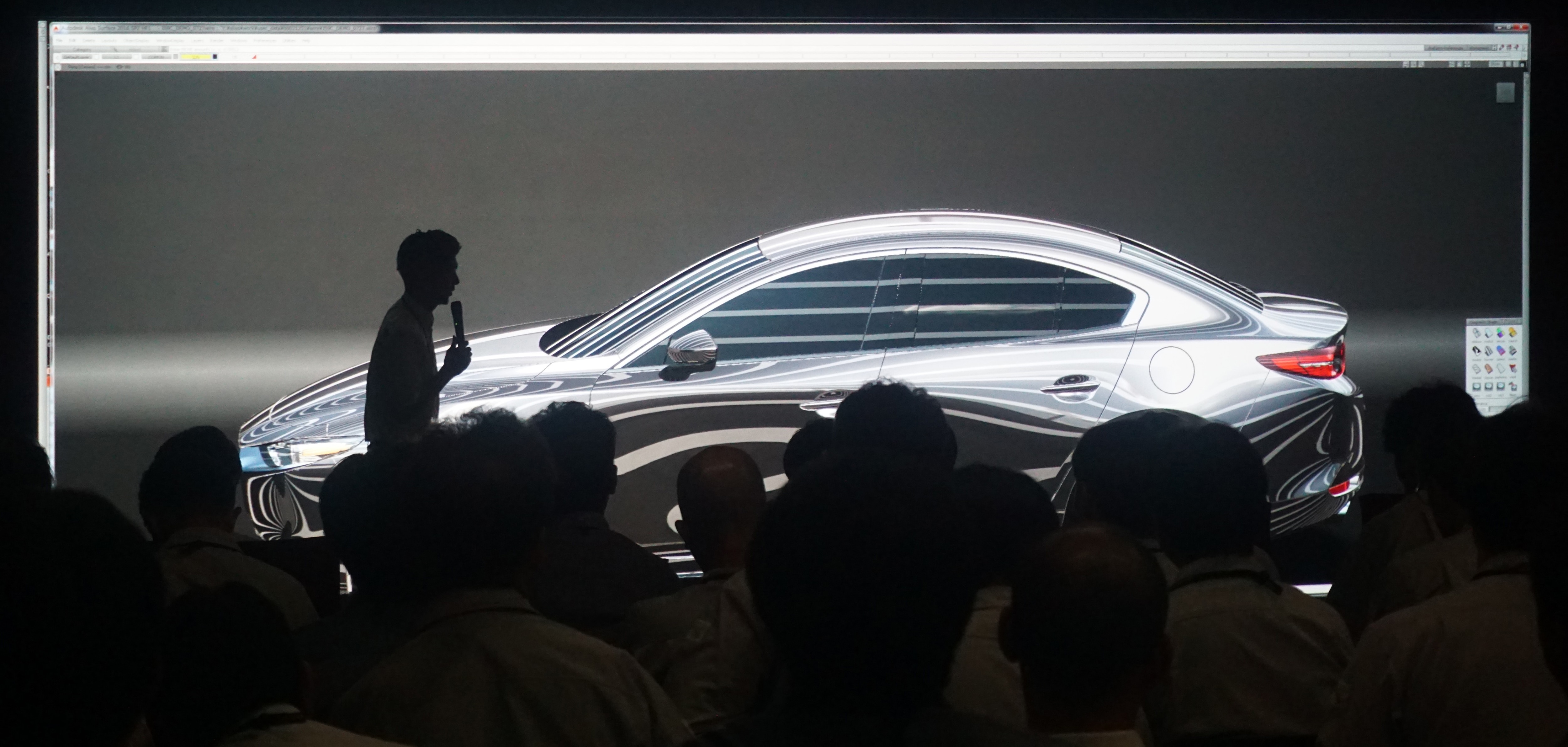 A presenter discusses a sleek car design displayed on a large screen to an audience in a dark room.