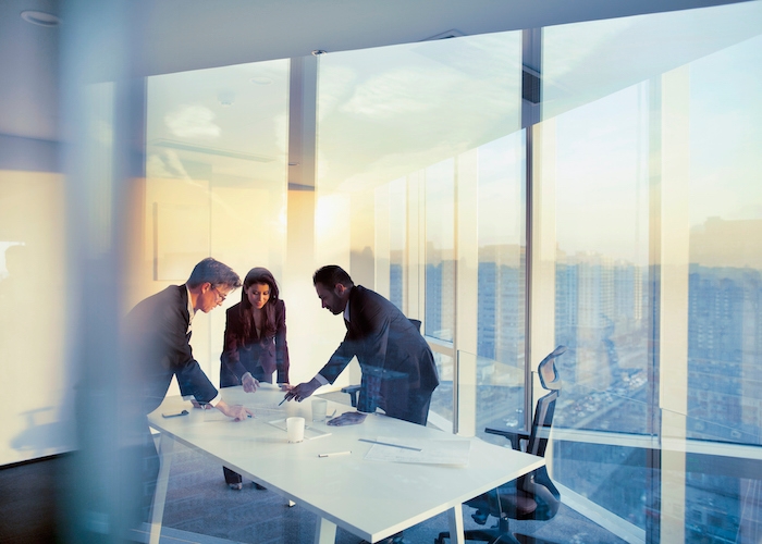 Two men and a woman stand around a desk in an office with floor-to-ceiling windows.