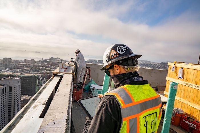 A construction worker consults a tablet as he stands on a building roof.