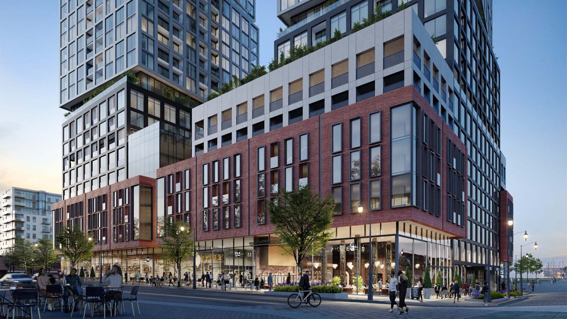 Modern mixed-use building with red brick facade, large windows, retail spaces, outdoor seating, and people walking and biking.