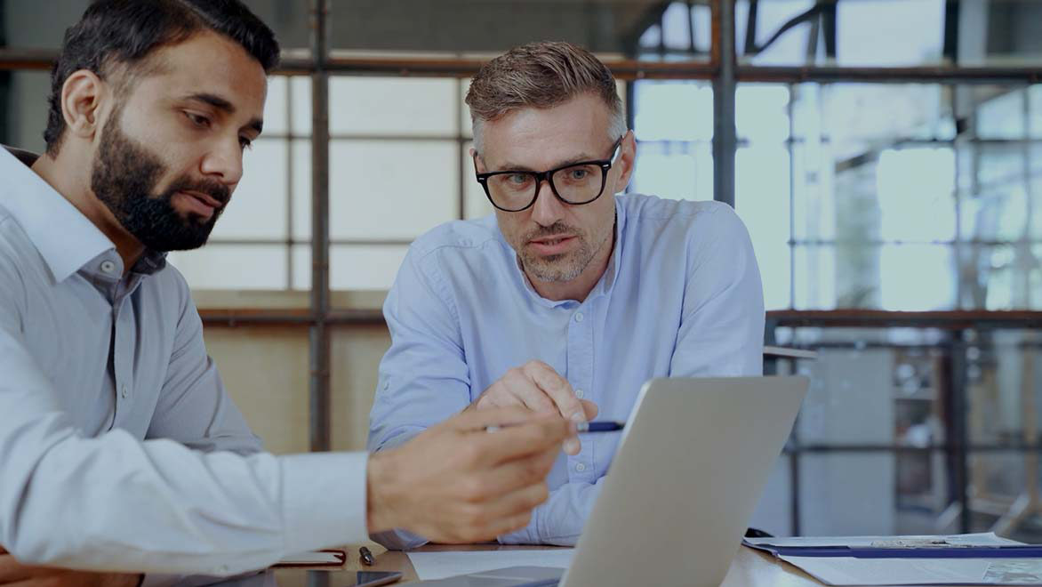 Two colleagues seated at a table review information on a laptop while discussing and pointing at the screen.