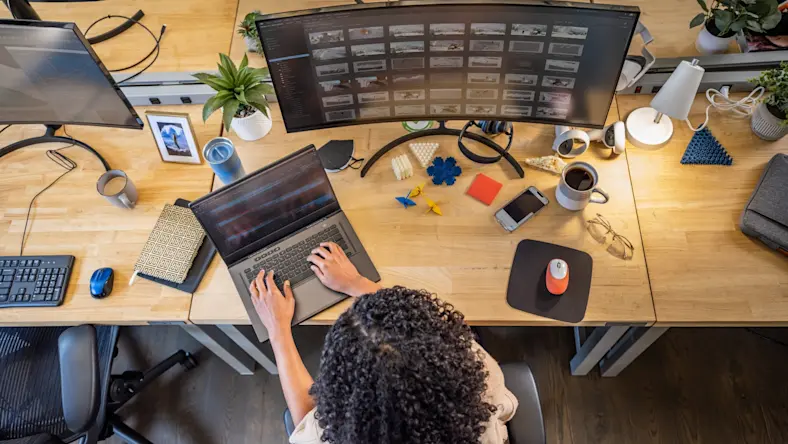 In an overhead view, a woman works on a laptop in front of a large curved monitor.