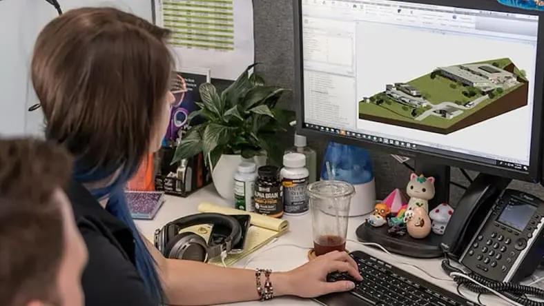 Over-the-right-shoulder view of two BHI employees (a man and a woman) sitting at a desk looking at an AutoCAD rendering on a computer monitor of Build Health International's Saint Rock Hospital project in Haiti.