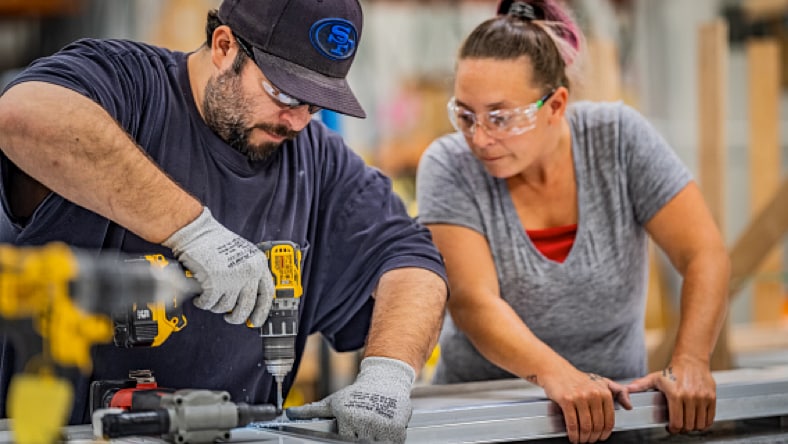 Two Pallet employees, a man and a woman, working on the Pallet production line. The man (left) is wearing a navy blue t-shirt and work gloves, using a power drill to drill a hole in a shelter component as the woman (right)—wearing a grey t-shirt and holdin