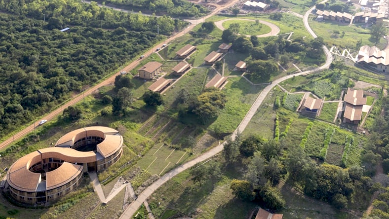 Aerial drone photo of the MASS RICA campus, with the main infinity-shaped building in the lower-left corner, with a series of buildings appearing intermittently on the campus grounds. The campus is surrounded by forest and includes what appear to be crop