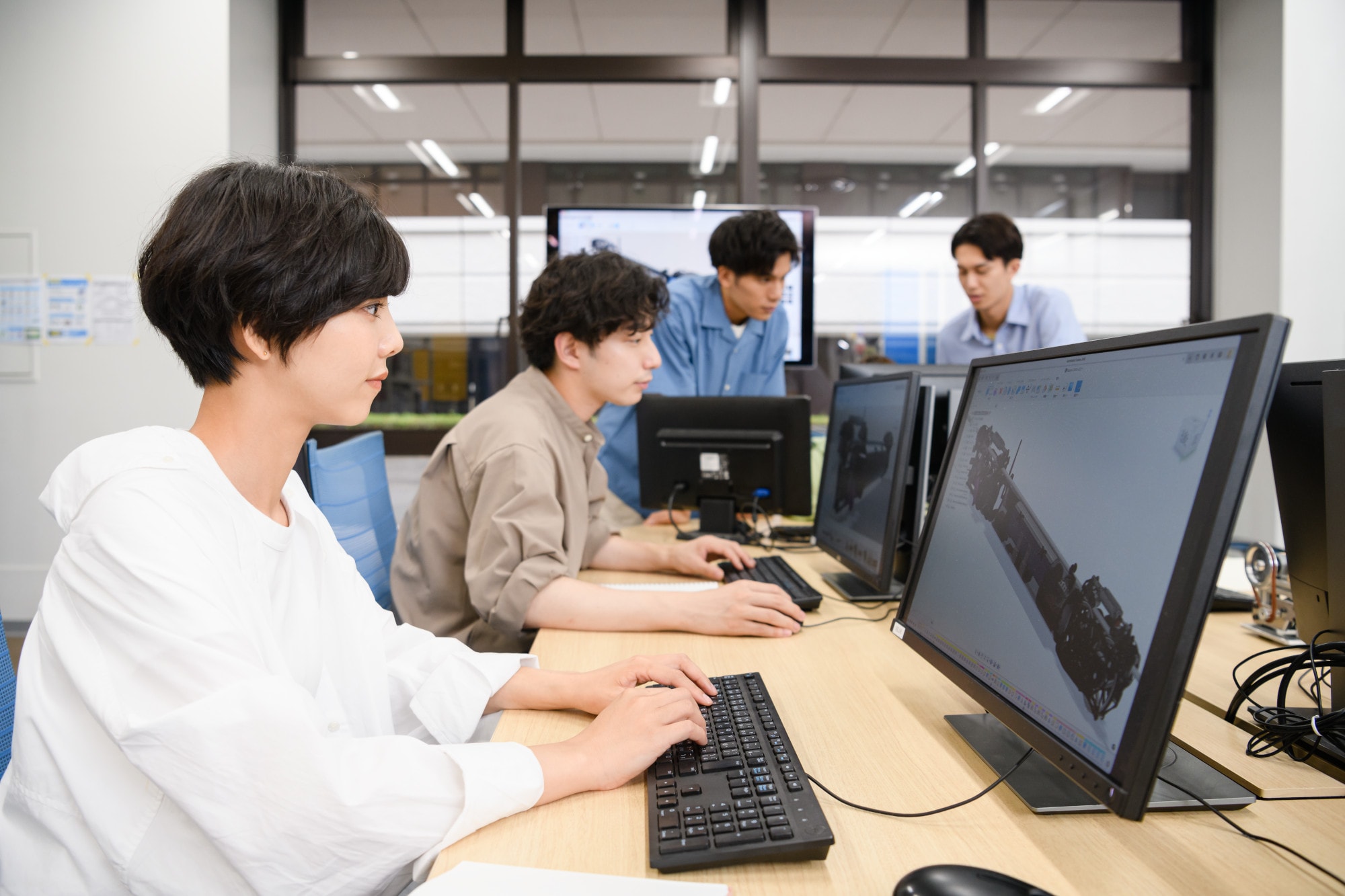 Four students work together in a computer lab, focused on desktop screens displaying a 3D design model.