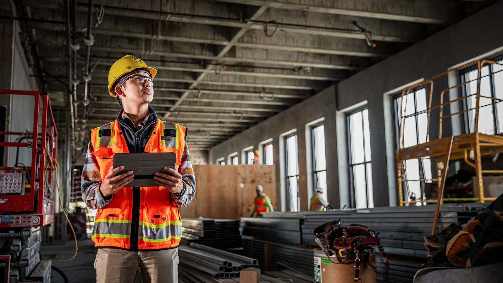 A construction worker stands in a large building, holding a tablet and reviewing plans or data.