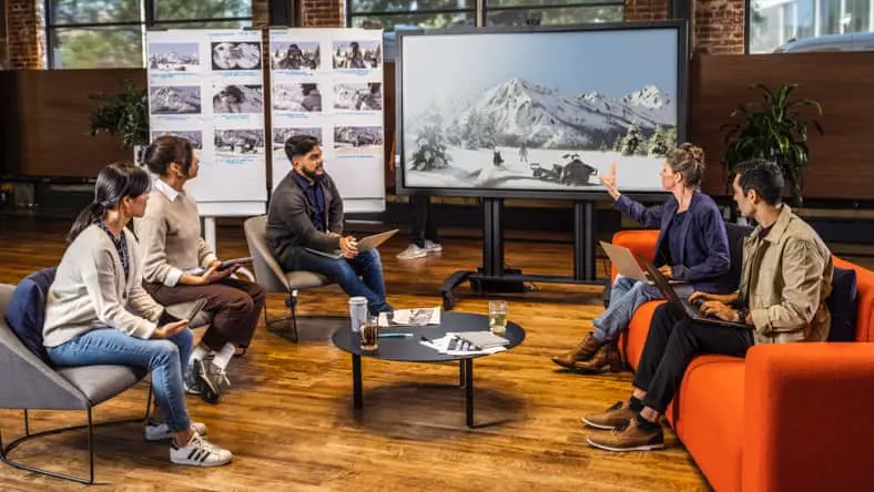 A group of five colleagues sitting in a workspace at separate chairs and couches looking at a large flat-screen TV with a snowy mountain and snowmobile displayed on the screen.