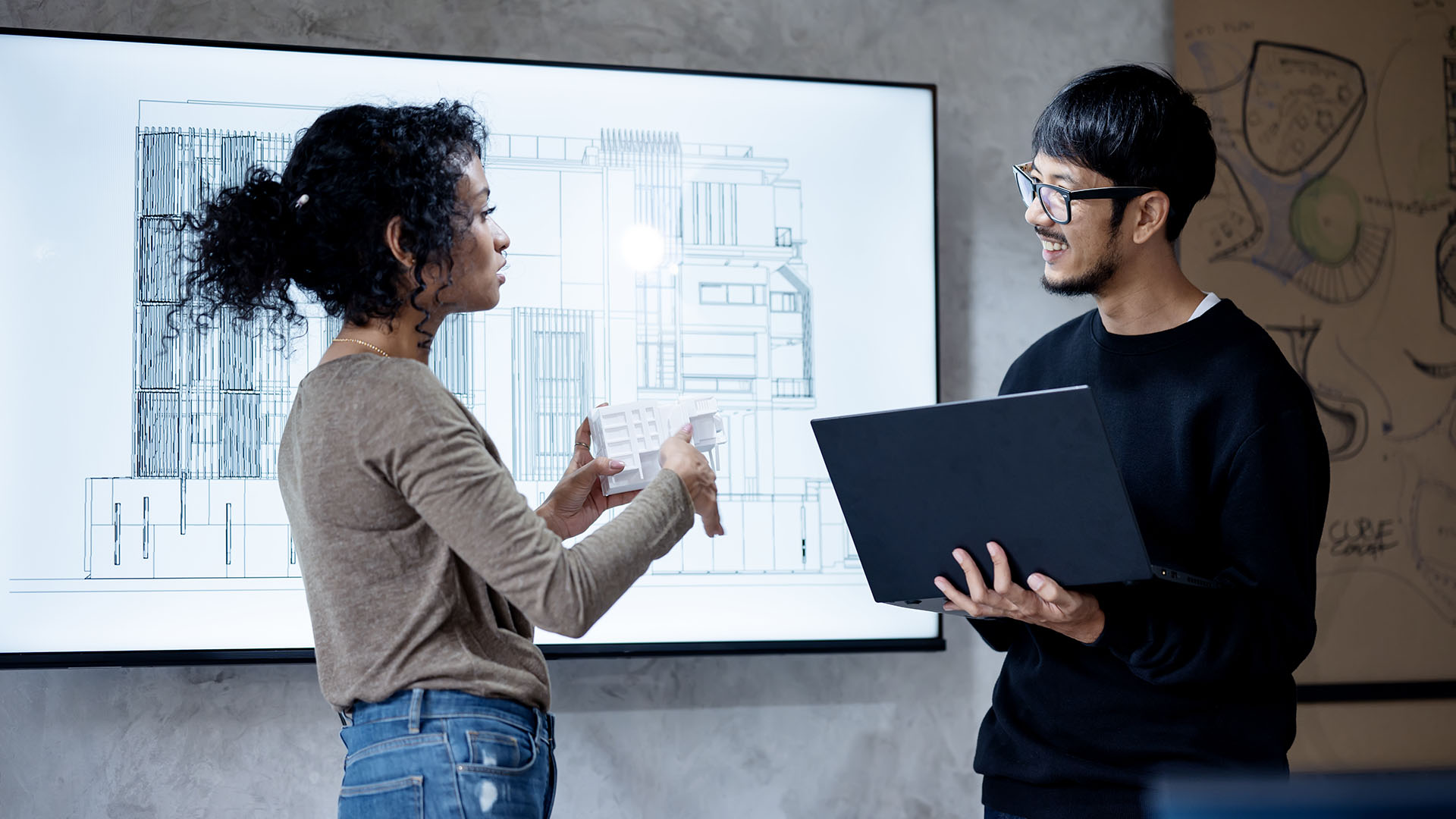 Two workers discuss a building model design on a large screen on the wall behind them.
