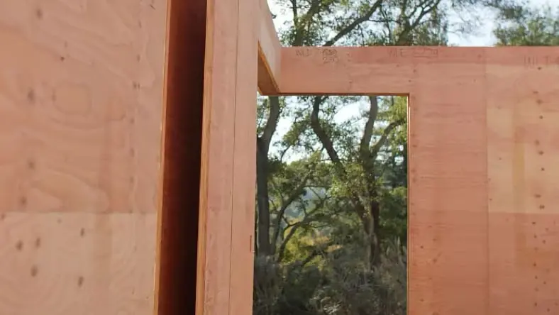Wide-angle view of the inside corner of a room under construction, with a large hole cut out for a door and no roof or ceiling, revealing sprawling trees in the background.