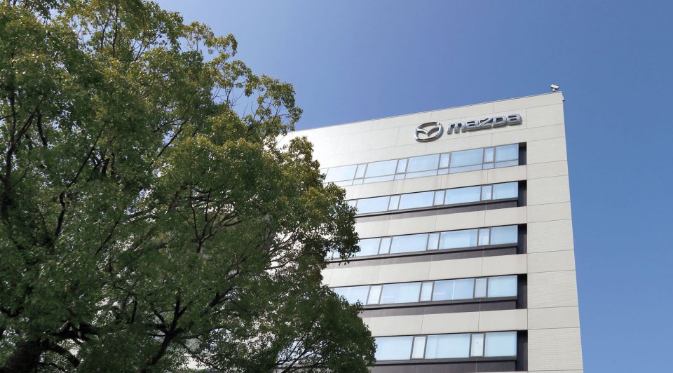 A modern building with the Mazda logo on top, surrounded by greenery under a clear blue sky.