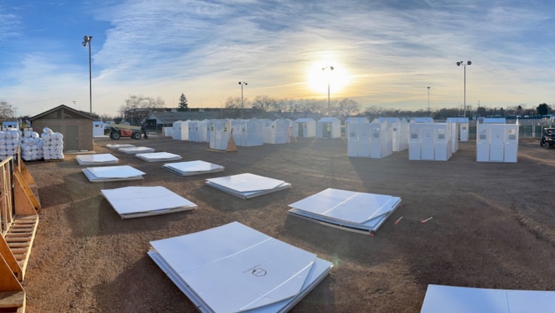 Wide angle view of a Pallet shelter village in development, with rows of standing shelters, nearly complete, in the background, and two more rows of flattened shelter components, organized into stacks, ready to be built, in the foreground.