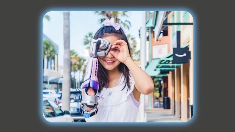 Young girl smiles while peering through imaginary binoculars using her hand and her Limbitless prosthetic arm.